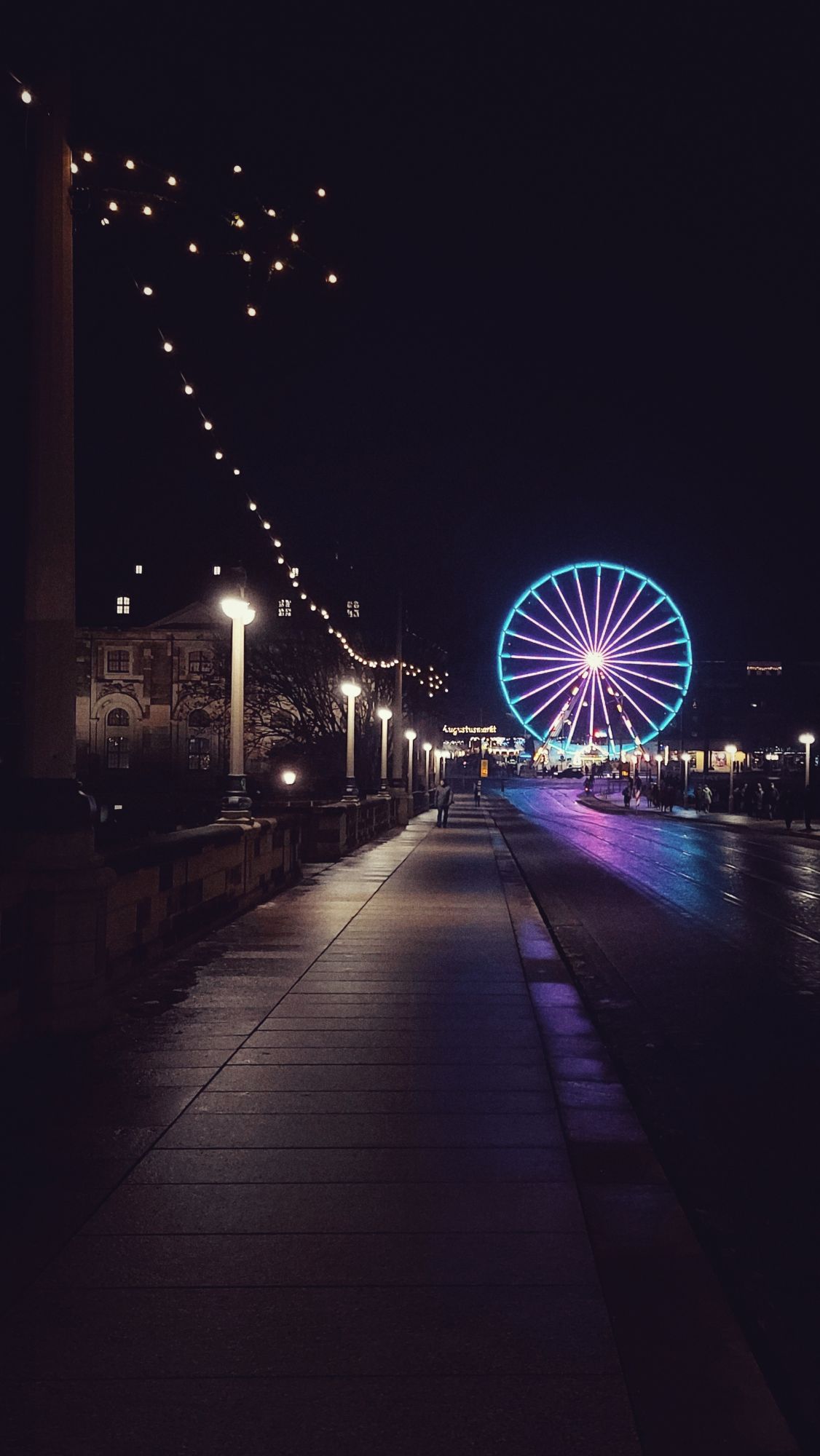 A coloured ferris wheel at the end of a street. Some featureless people. Night scene.