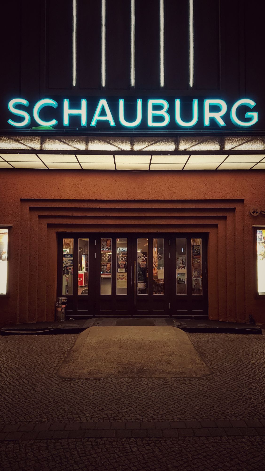 Front of a movie theater, with closed doors and an empty entrance. A bright blue neon logo reading SCHAUBURG.