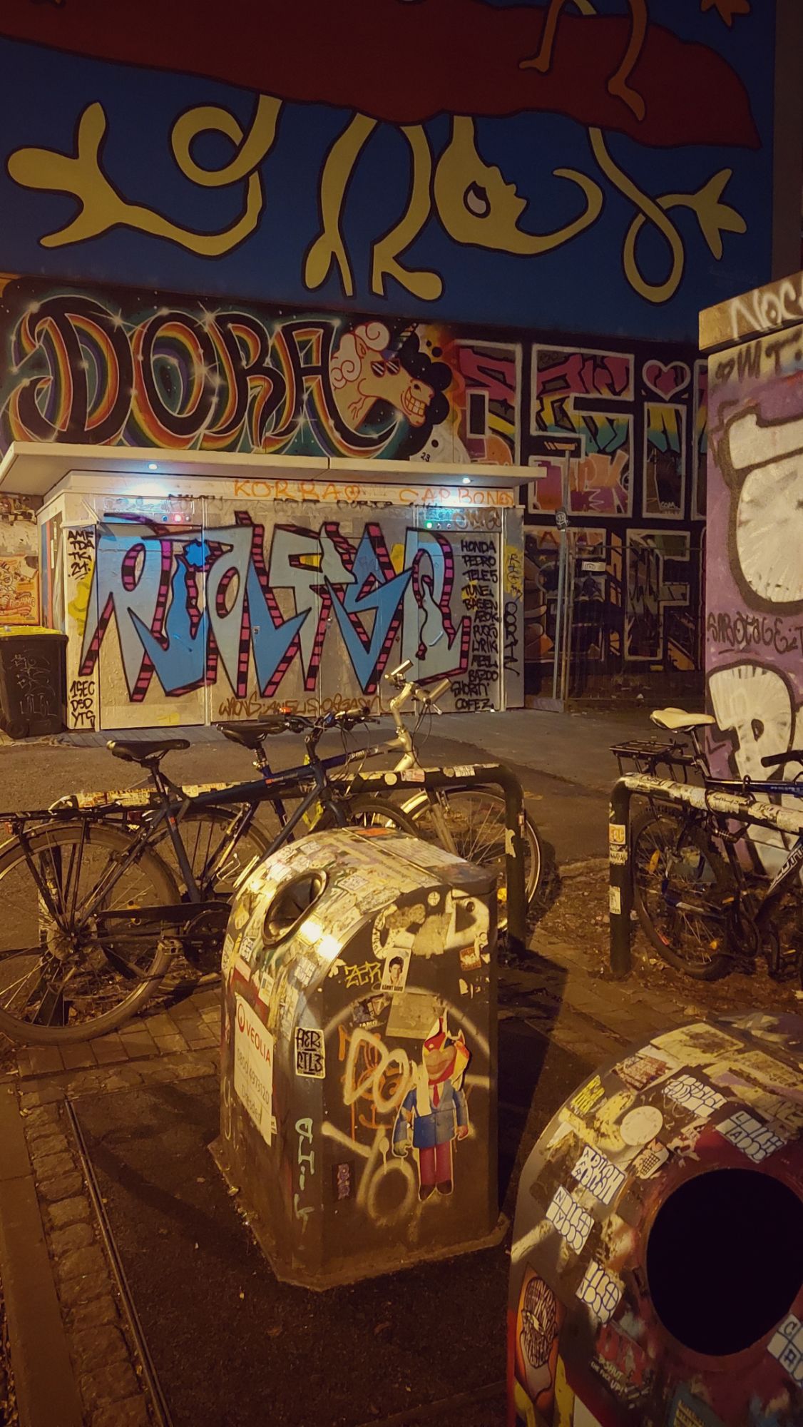 Street scene: A graffiti-laden wall, bicycles tied to iron bars, and garbage cans in the foreground, stickered over and over.
