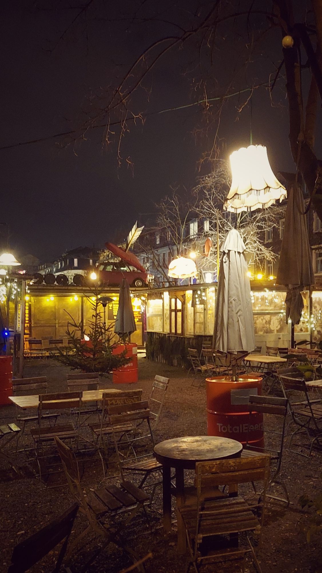 View into an outside bar. Bright lights, a car and living room lamps decorating the scenery. Closed umbrellas, wooden tables and chairs. The location is closed.