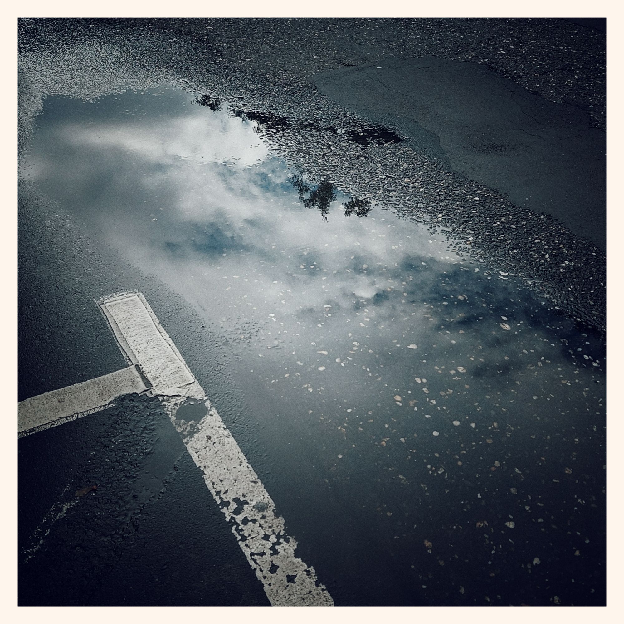 Reflections of clouds and blue sky on a flat puddle of water. Guiding lines of a parking lot left of it.