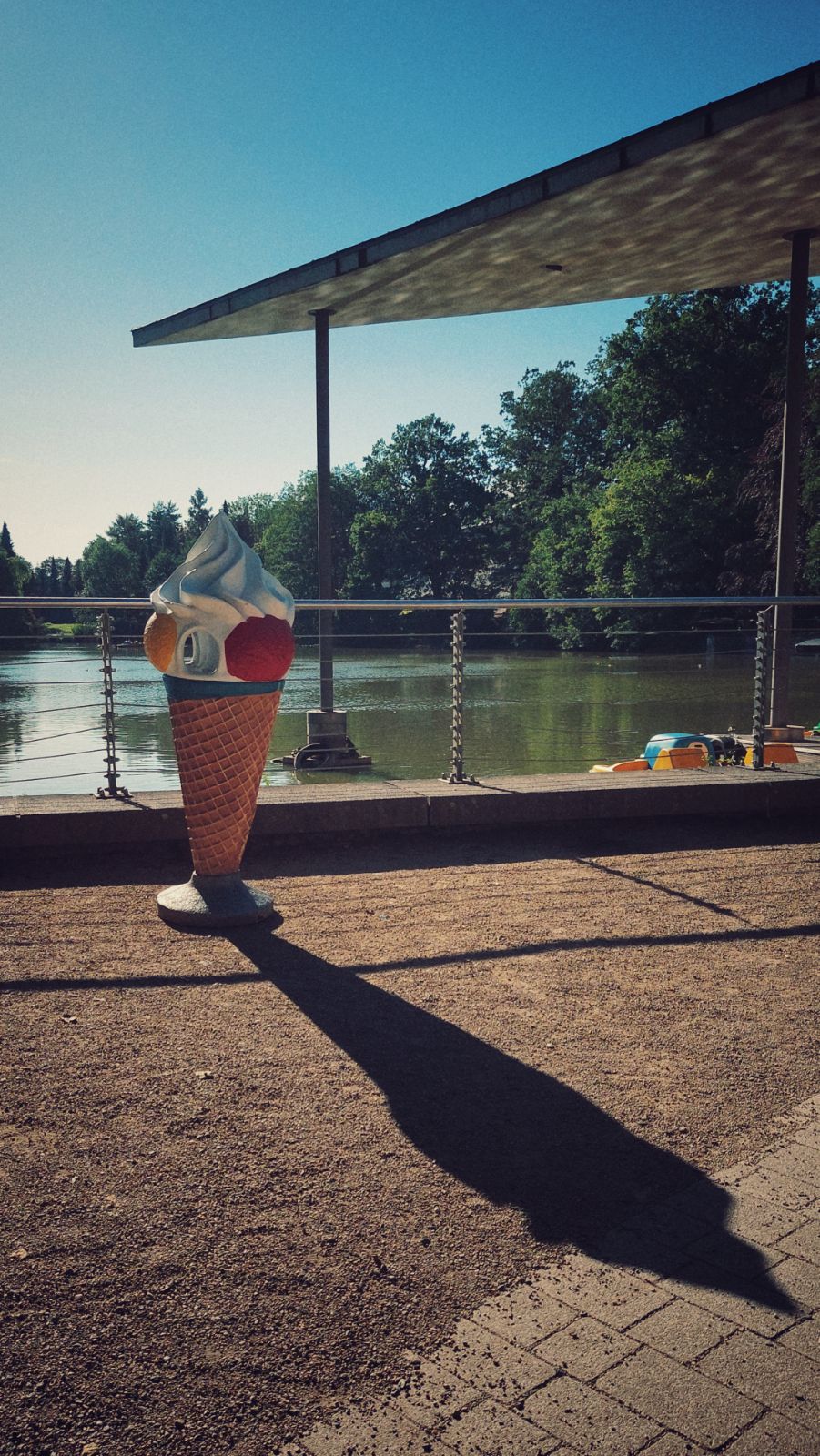 A large ice cream cone casting a long shadow next to a lake. There are small boats on the lake and a flat roof above.