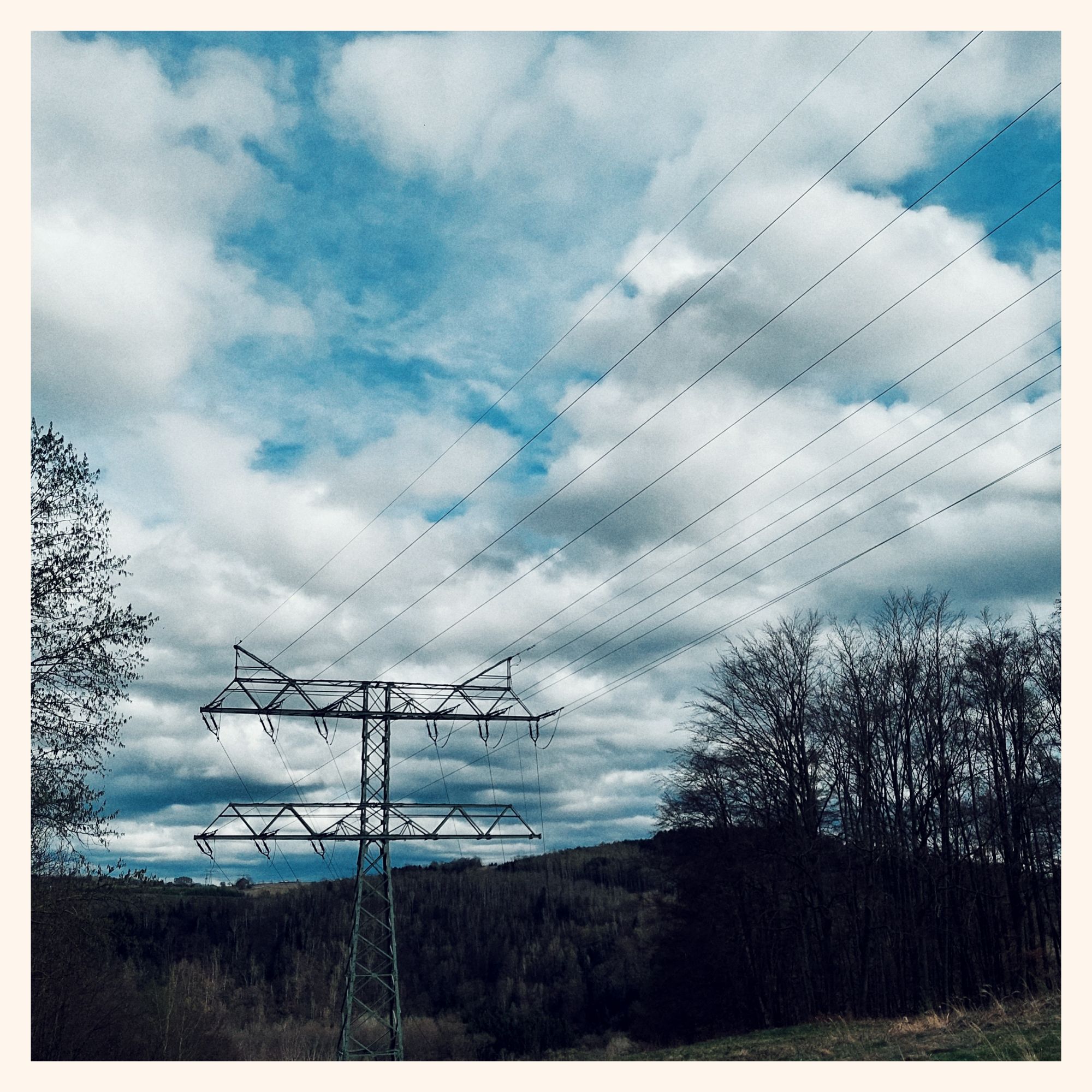 Looking along a power line in a forest. Blue sky, some large clouds, trees, dark hills in the lower part. Reasonably warm colors.