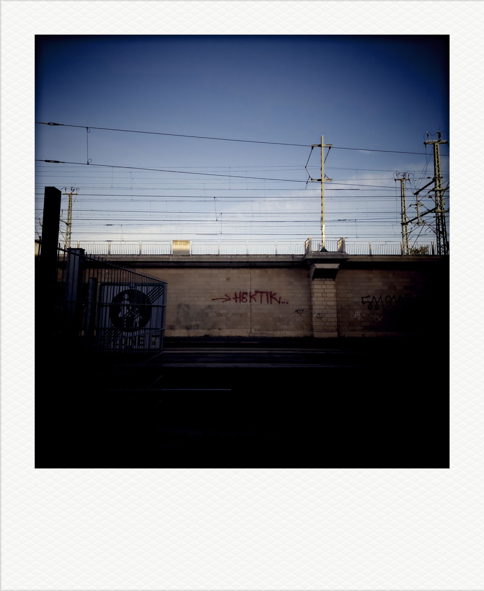 Straßenszene unter blauem Himmel. Bahndamm, Oberleitungen, eine Mauer, ein rotes Grafitto: "Hektik".