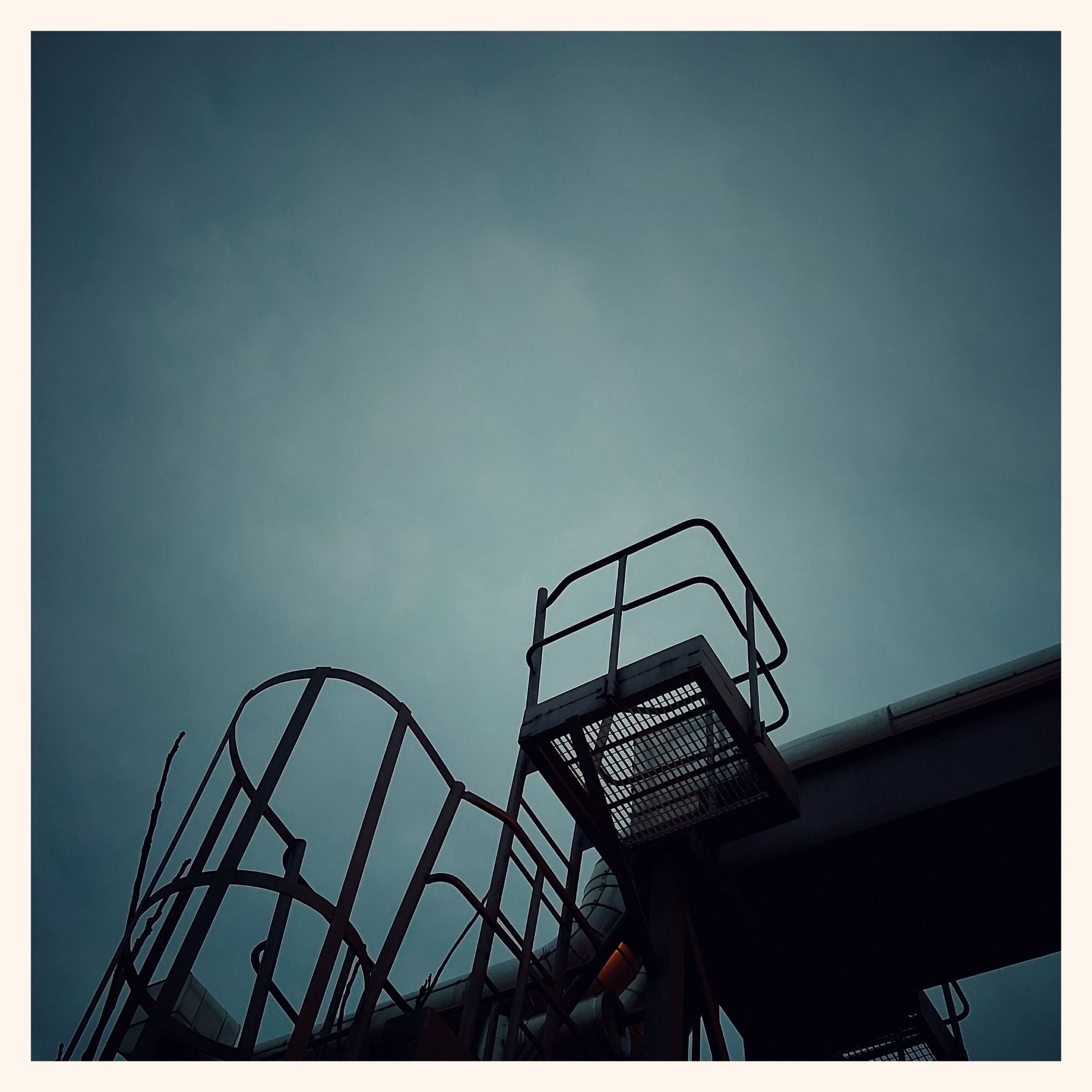 Grey sky above industrial construction. A ladder leading up to a steel bridge carrying pipes.