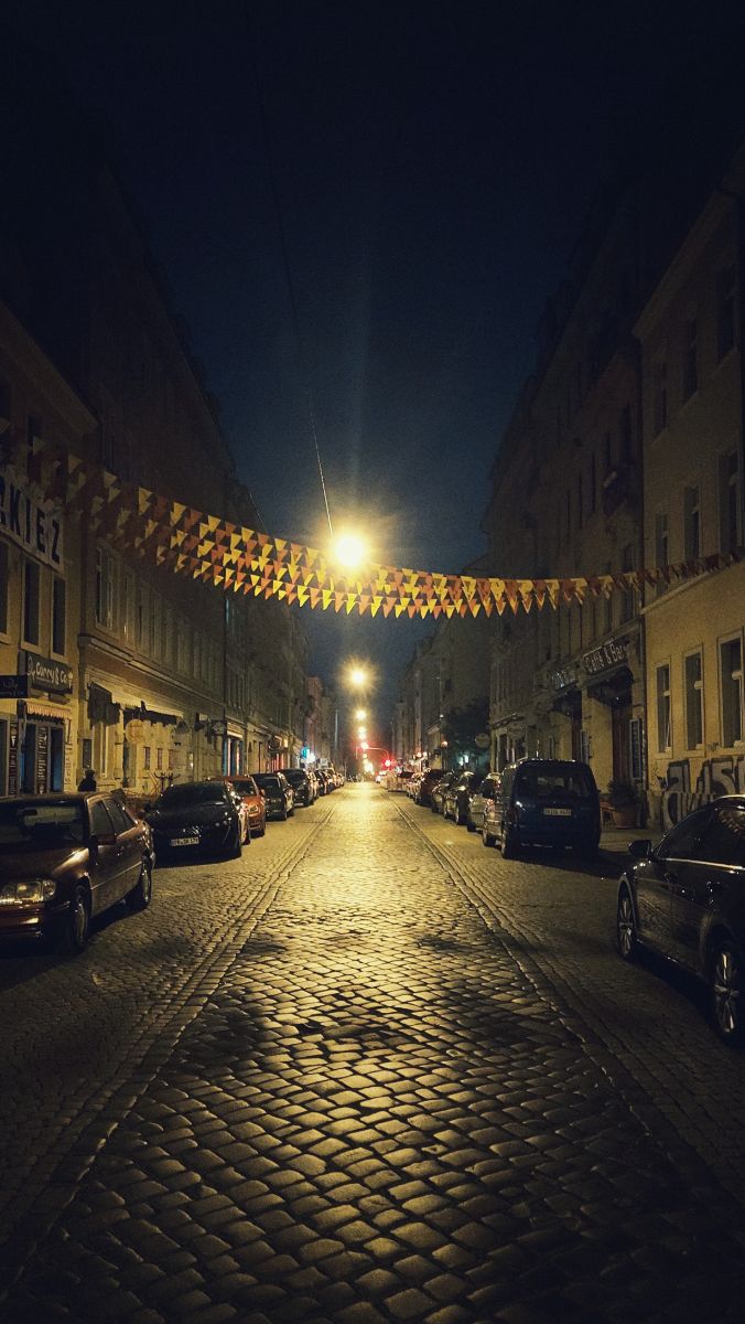 An empty city street at night. Old facades, parking cars, small flags in artificial light.
