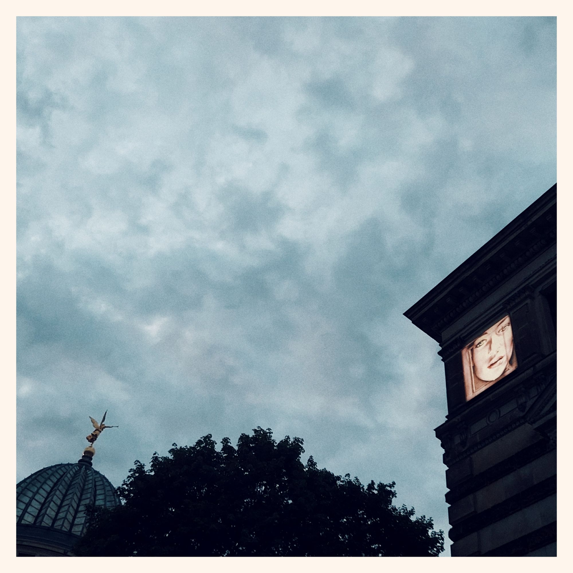 A sea of grey clouds, below an old town silhouette: A glass roof resembling a lemon squeezer with a golden statue on top. Dark trees. And a corner of an old house with a lit painting on it 