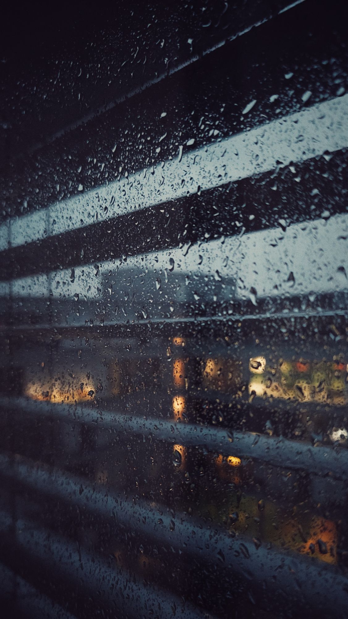 Window blinds and office lights behind rainy windowpane.