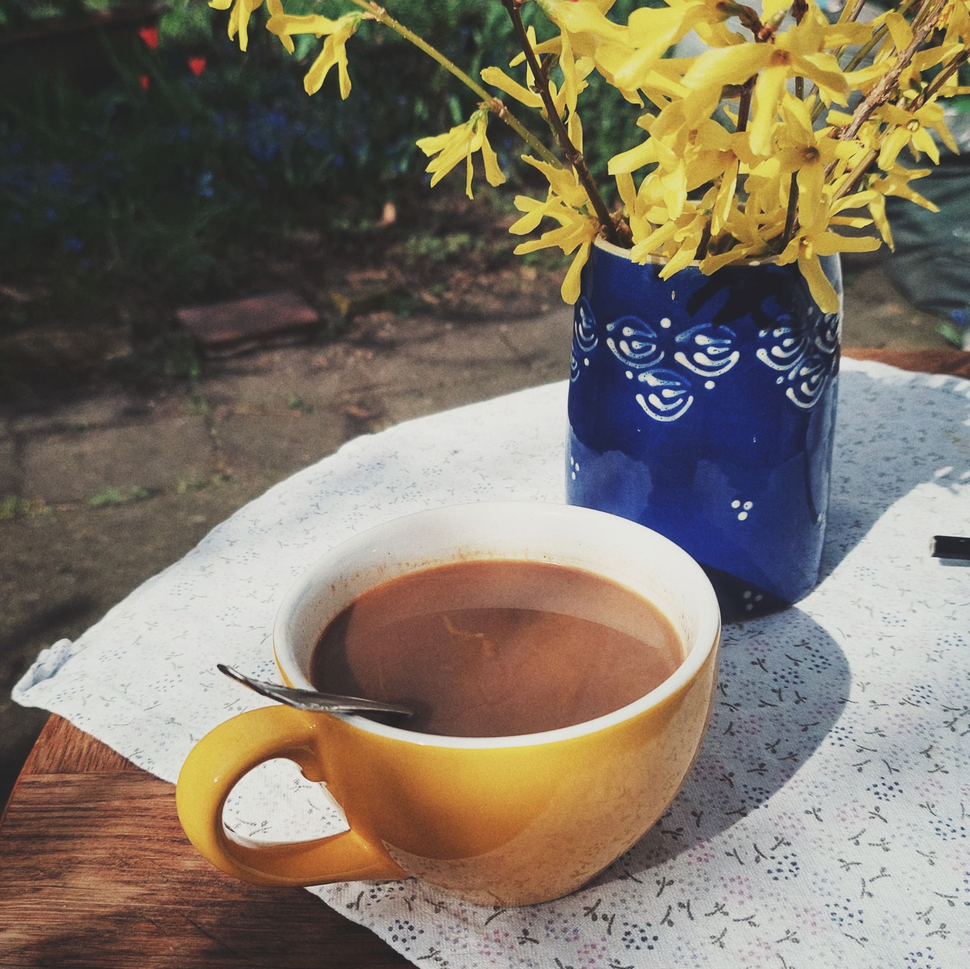 A yellow cup containing coffee on a wooden table. Forsythia branches in a blue vessel.