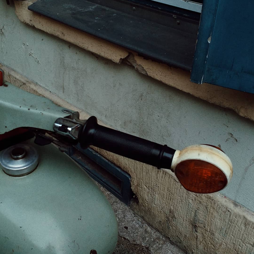 Part of an old motorcycle, a steering lever and a direction light in front. In the back, wall of an older house, a blue window frame and a light green segment of concrete. Colour of the concrete and of the motorcycle seem to somehow match.