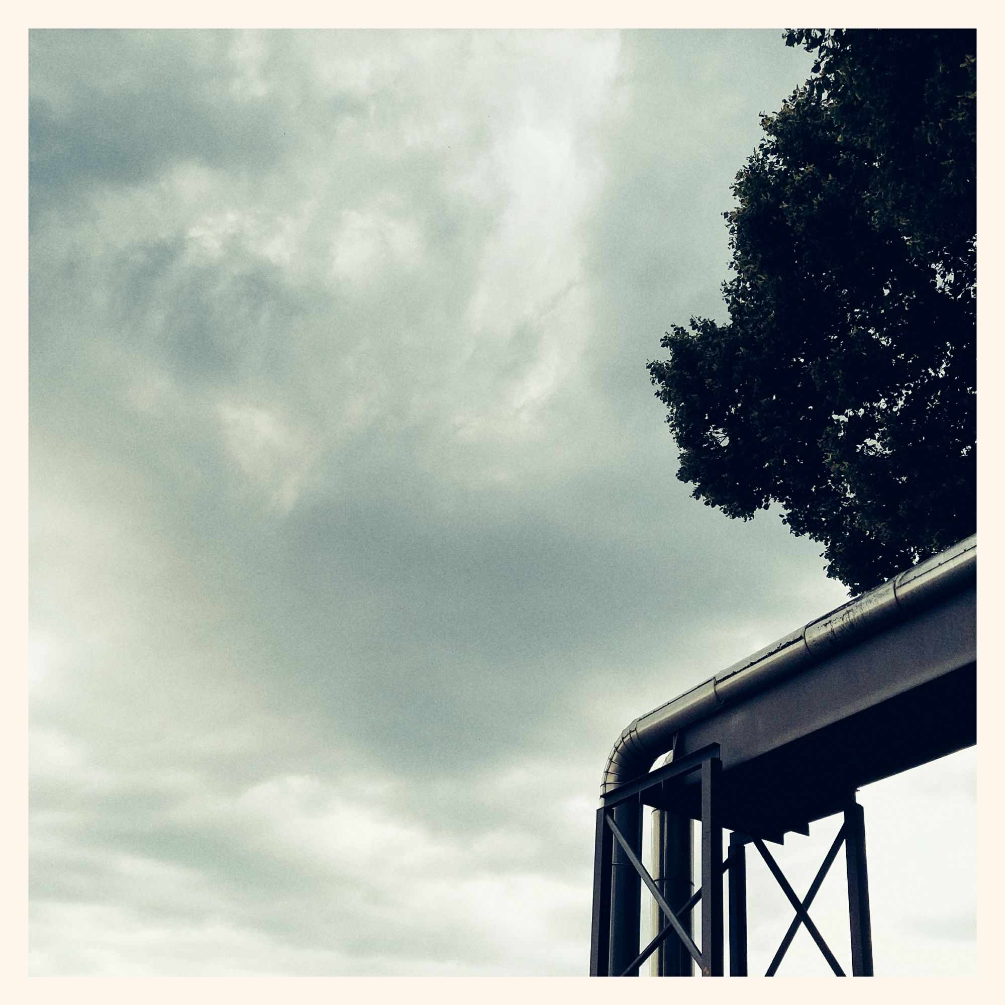 A tree on the right side of the picture, and the end segment of a bridge leading heat pipes across the street. Dense grey clouds on a light sky filling the remainder of the frame. Square cut, thin white border.