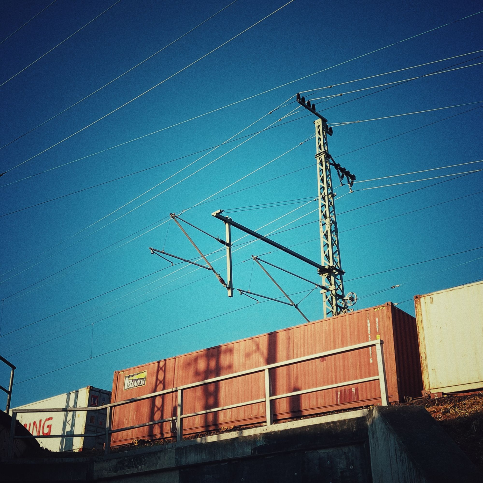 Blue sky, power pylons and lines. A train running behind the railings of a bridge.