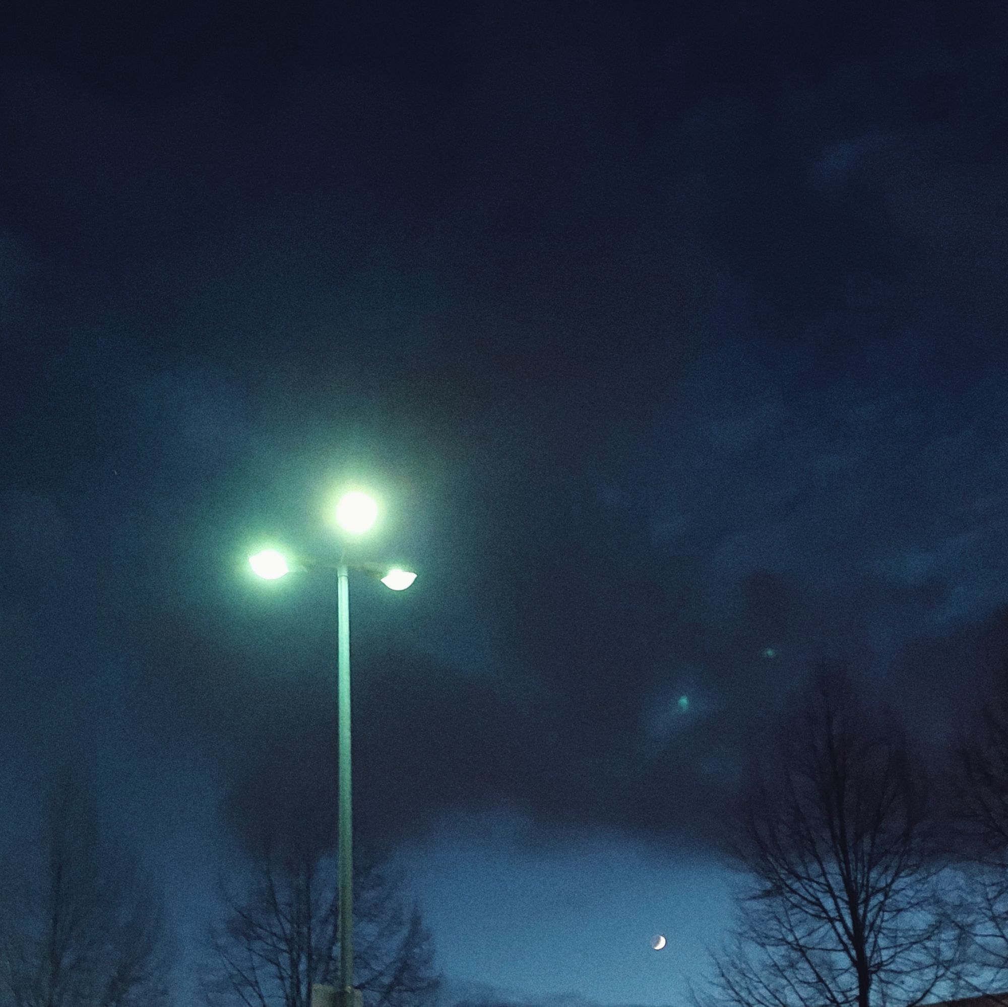 Sichelmond in später Dämmerung. Wolken, Stadtleuchte mit mehreren Flammen im Vordergrund. Flachdach darunter.