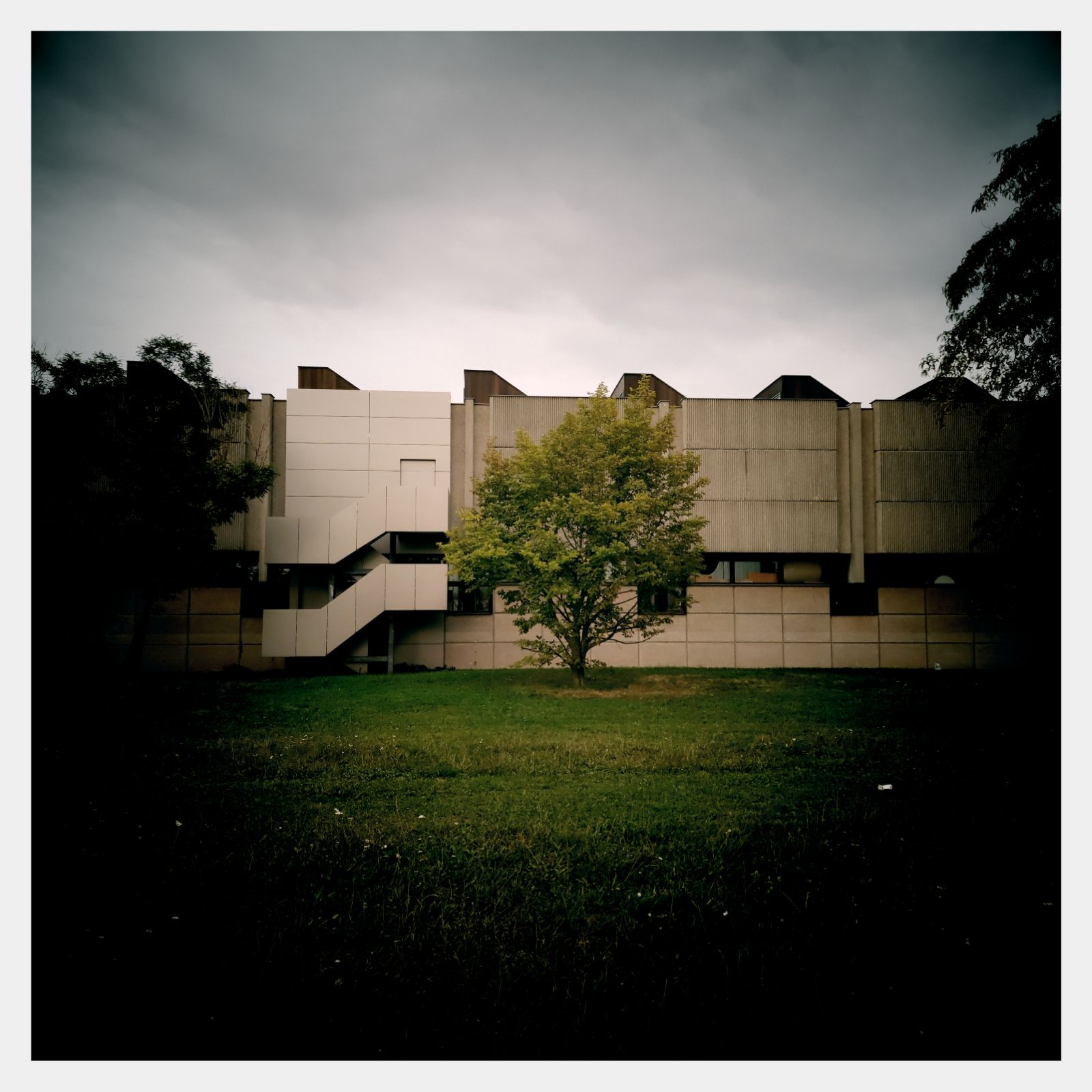 A socialist modern building facade, under grey skies, surrounded by trees.