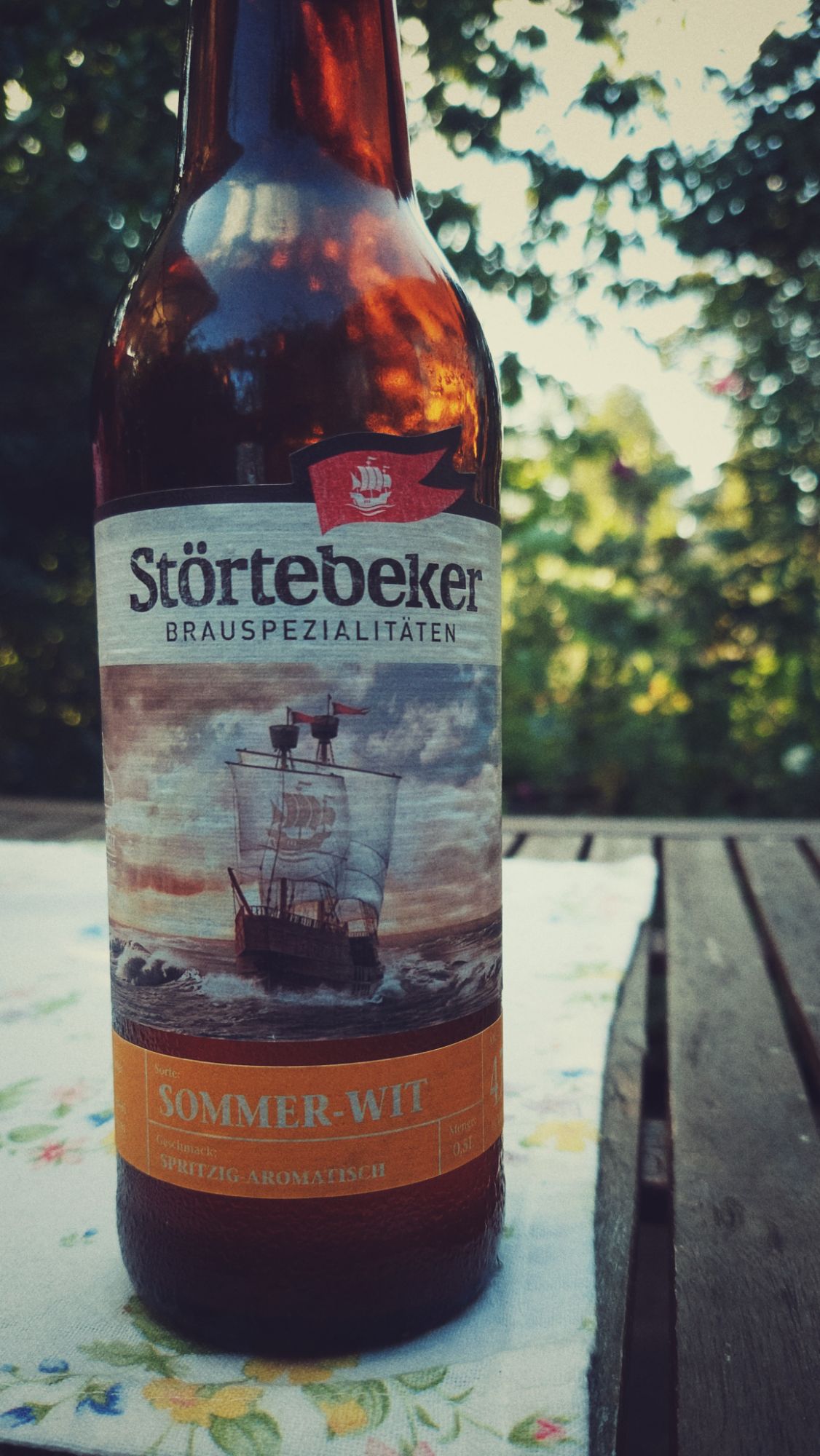 A bottle of Störtebeker Sommer Wit on a garden table. The tablecloth is white and features small flowers.