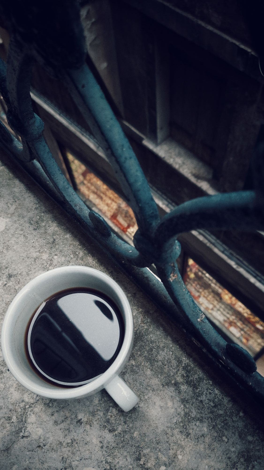 A mug containing black coffee, on a stone windowpane. Behind there's an iron railing, looking down into a small road and the storefront of a candy shop.