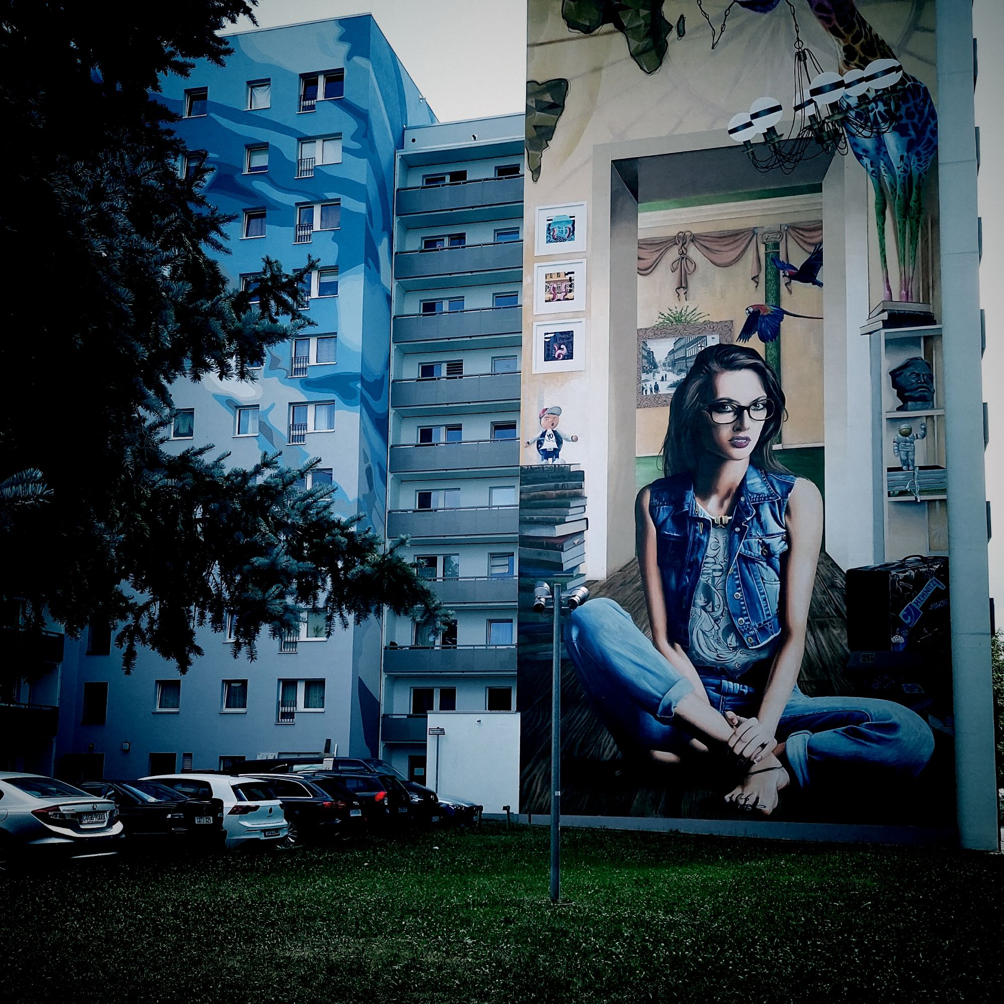 A mural of a young woman sitting in her small room, looking right at the spectators. Painted on the wall of a concrete housing block.