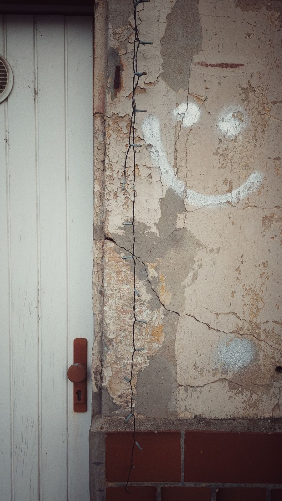Wall of an abandoned house. A white door on the left, a crumbling wall on the right. A white face sprayed to the wall, and a chain of small lights hanging down in the middle. 
