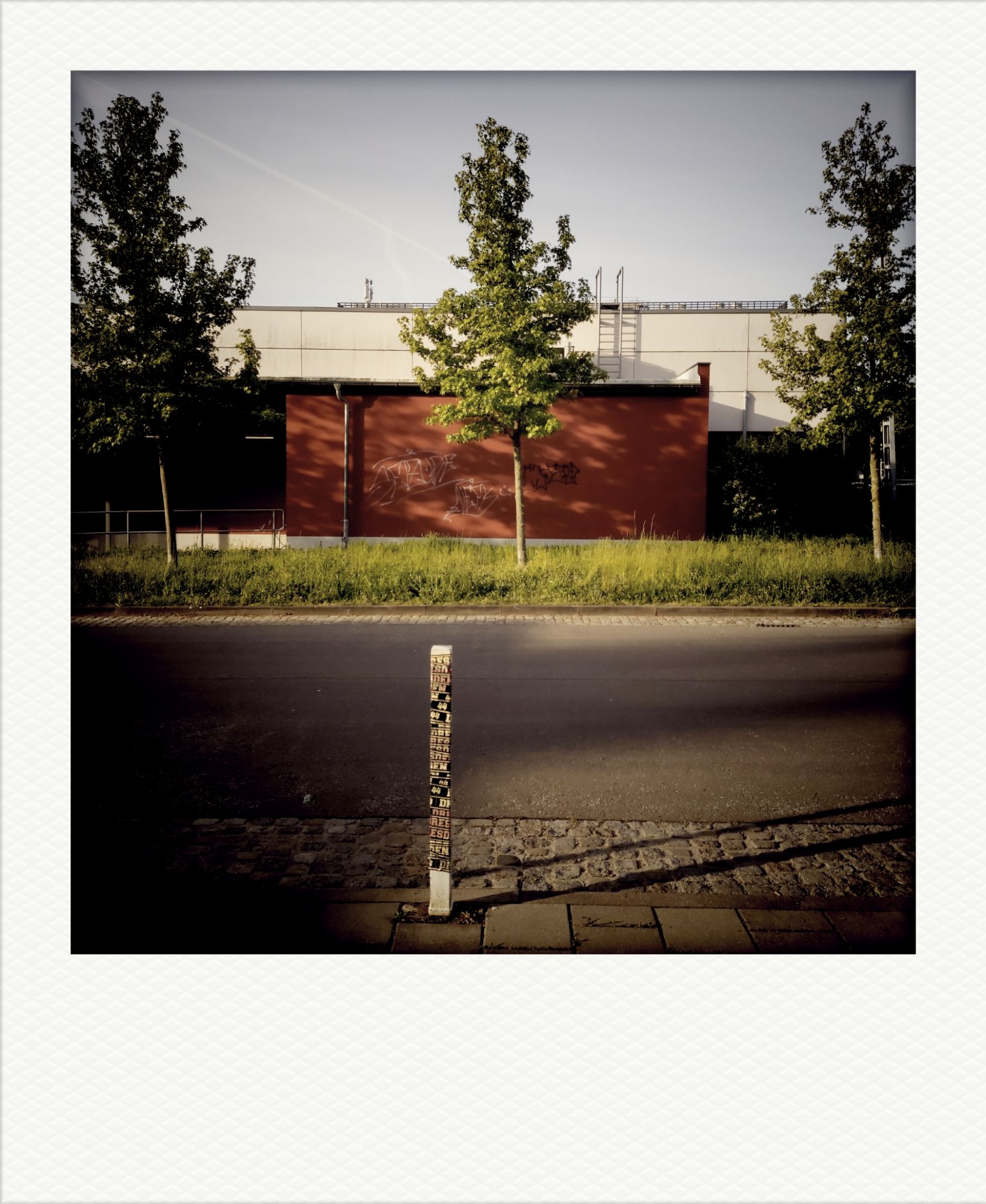 Stadtszene: Rote, weiß getaggte Wand. Wiese davor, ein moderner Neubau dahinter. Asphaltstraße, Fußweg, ein Boller übersät mit Aufklebern.