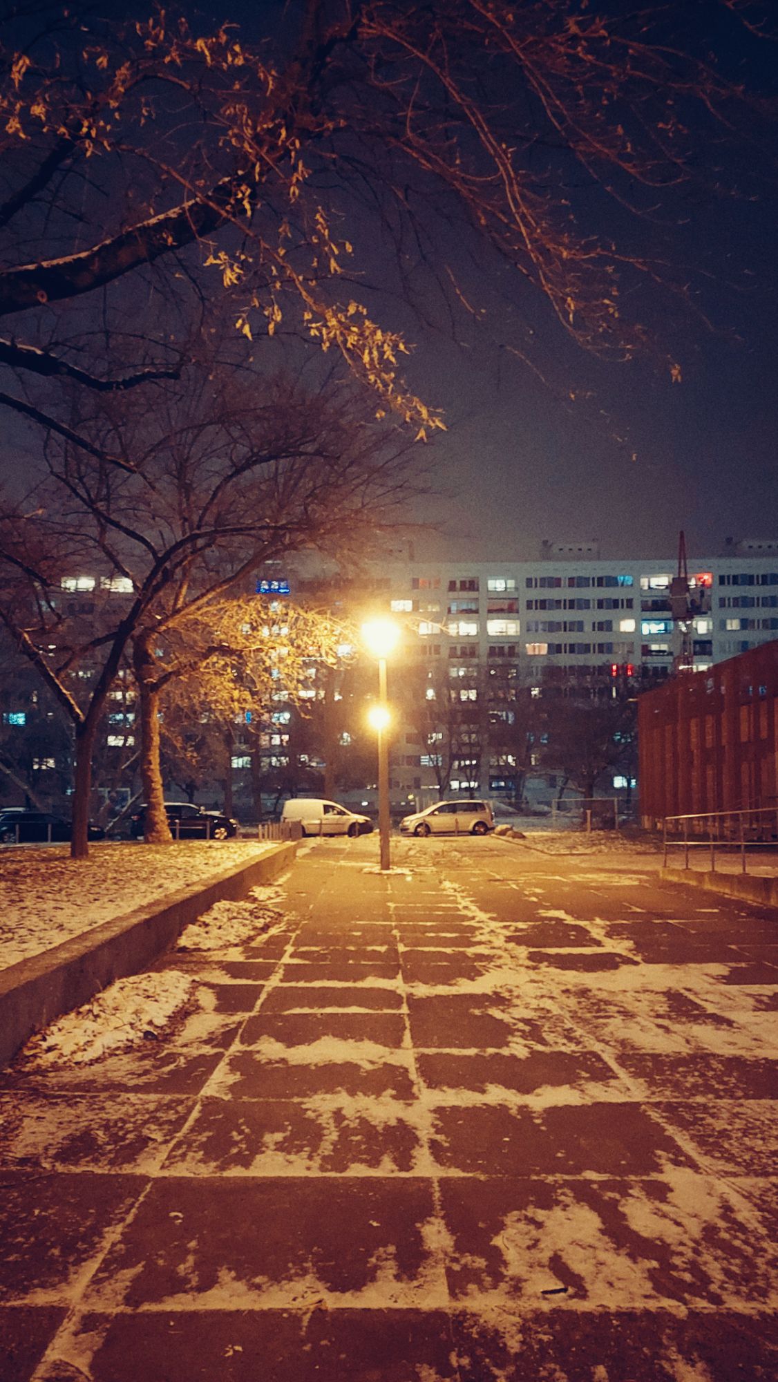 An empty passage between houses. Streetlights in a line. Some snow on the ground. Distant high-rises, and some parked cars.