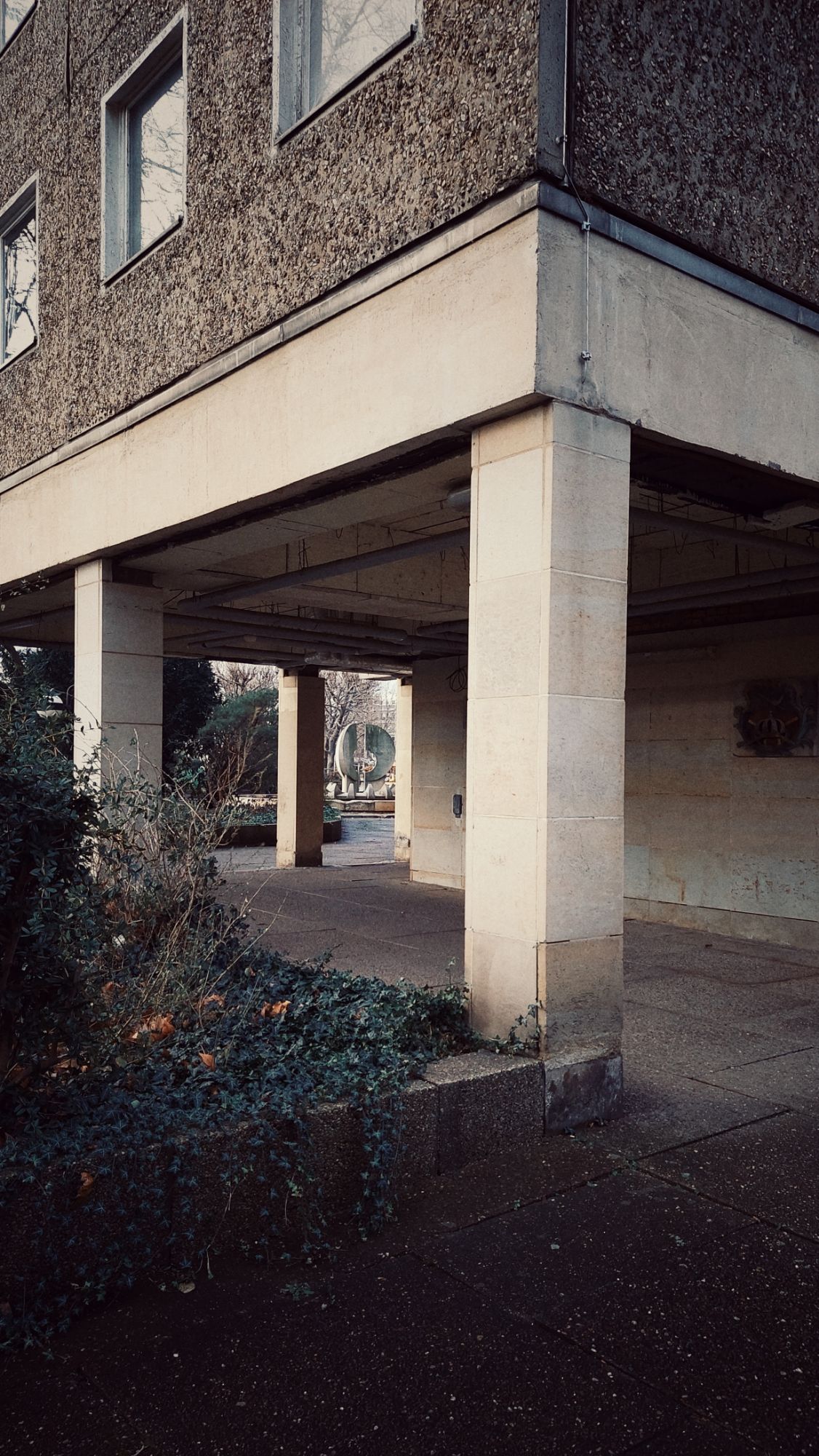 Concrete pillars as part of a building, with a partially open ceiling revealing pipes and other infrastructure. Left, a small piece of city green.