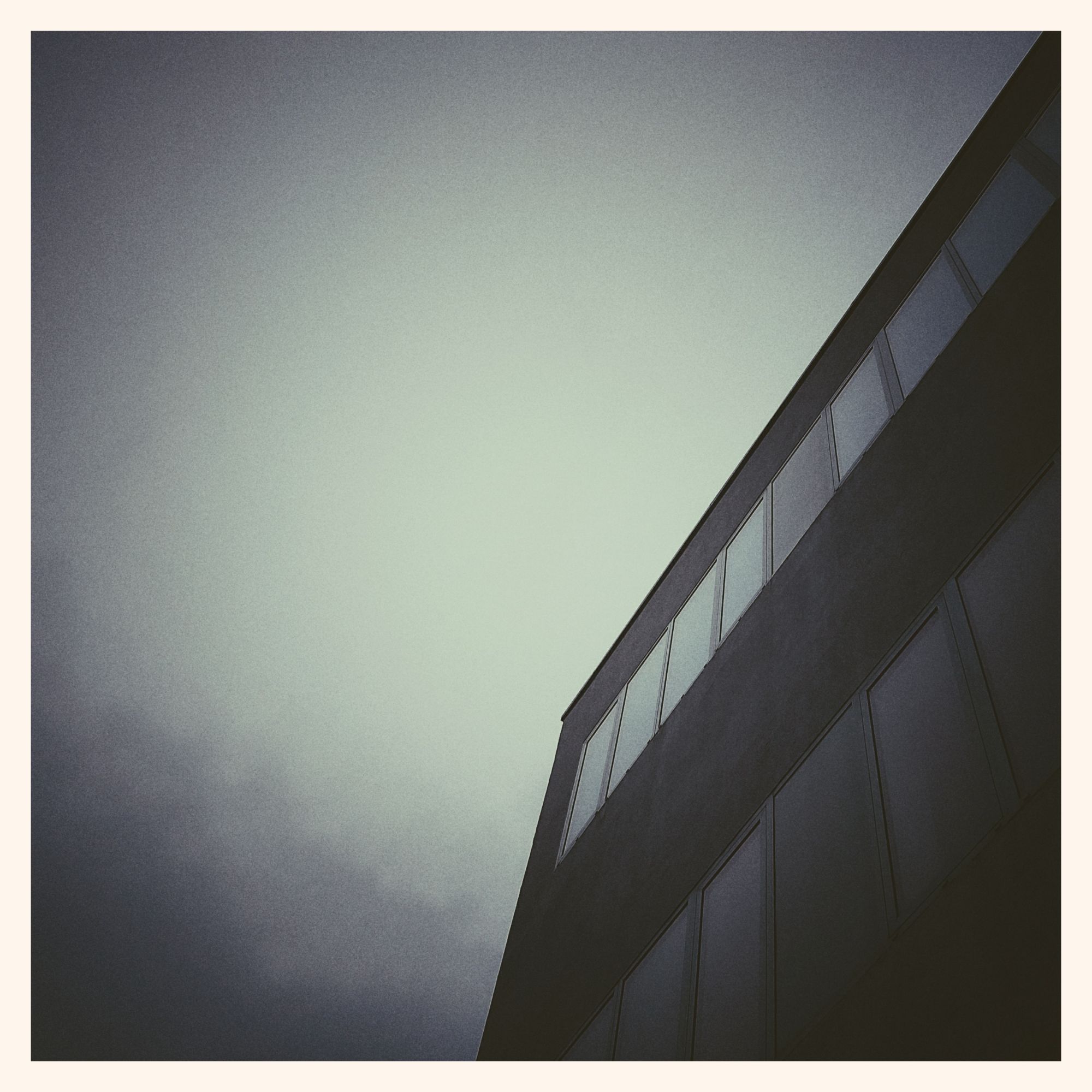 Pale skies, no clouds, above a modern-style building in the right part of the picture. Strong shapes, light reflecting in a line of windows. Old film effect.
