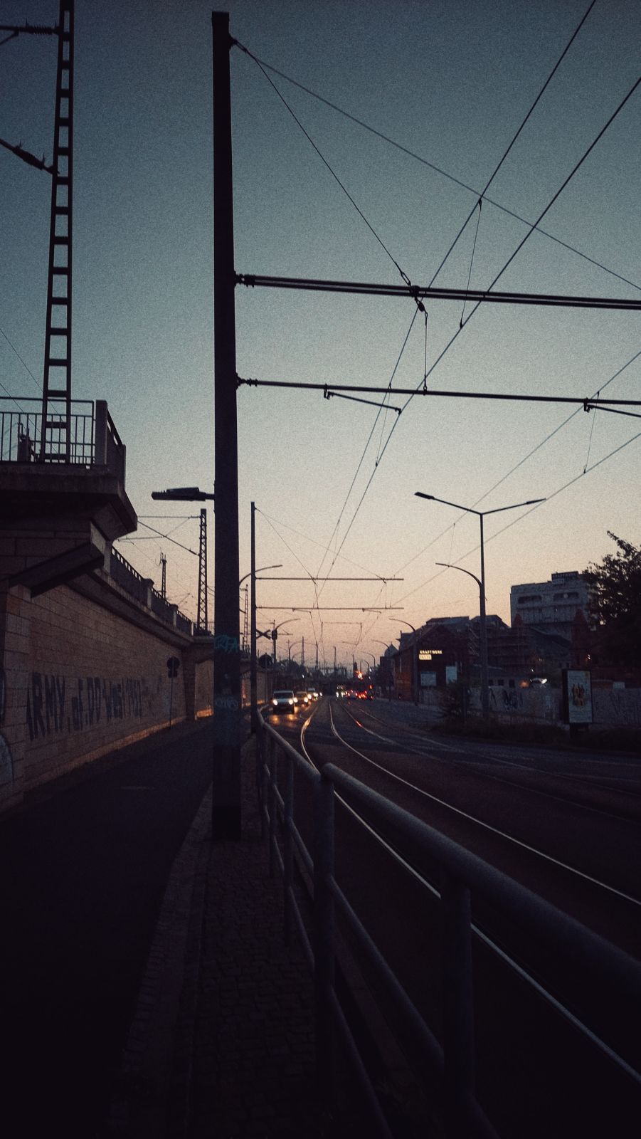 An early morning street scene. Tramway rails in front, between sidewalk and road. Some cars headlights approaching. Distant sunrise.