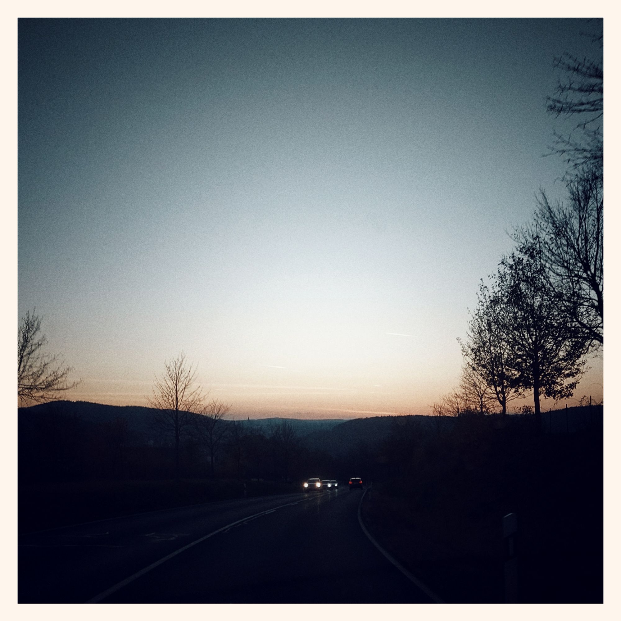 Pastel evening sky above hills and a street. Some car headlights moving in from the other end.