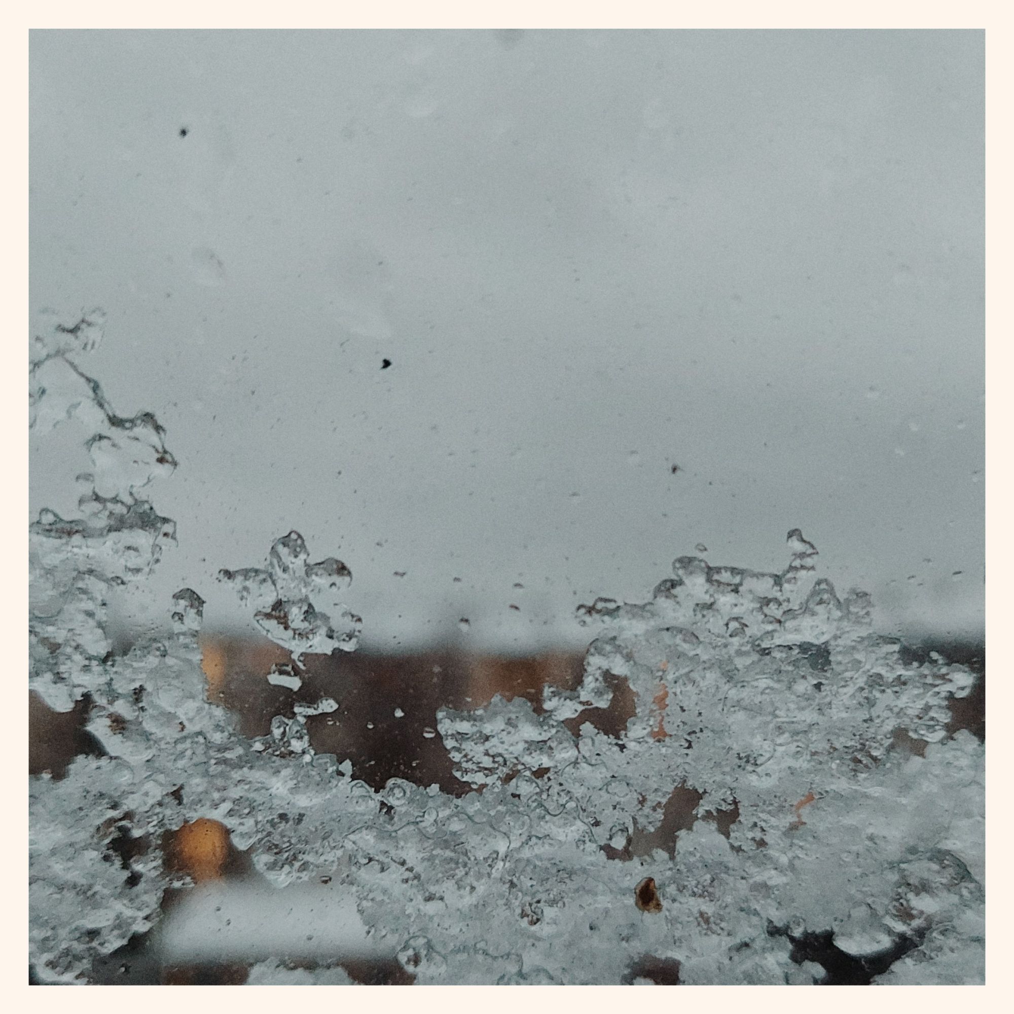 Frozen snow on a windowpane. Grey city sky and some houses with lit windows behind.