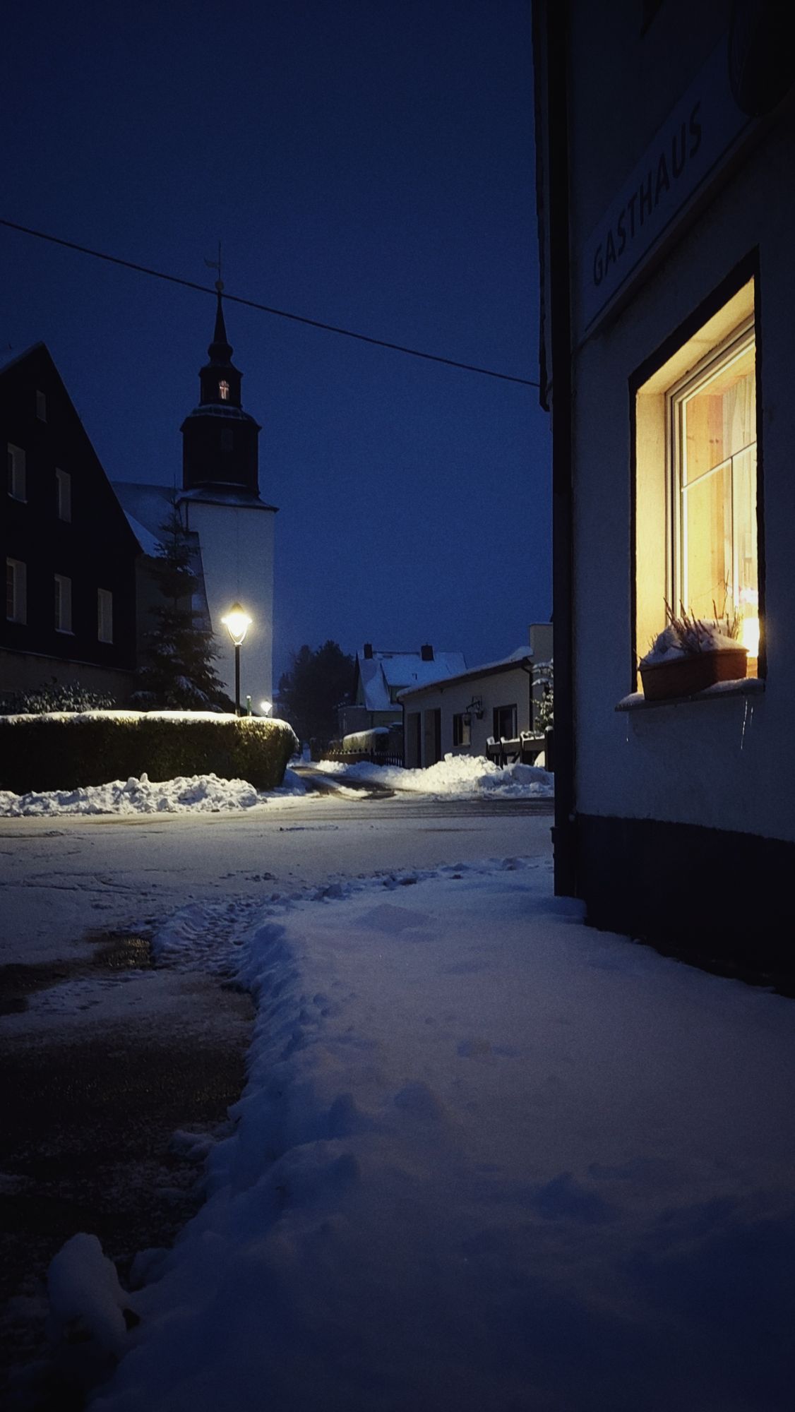 An empty village street at dusk. Warm light in a window, snow on the streets. Church and belfry in the back.