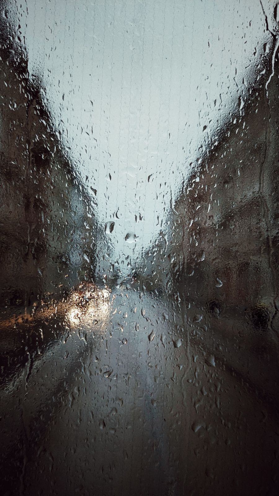 A city street seen through rainy windows. Headlights of a few cars to the left. Houses on both sides.
