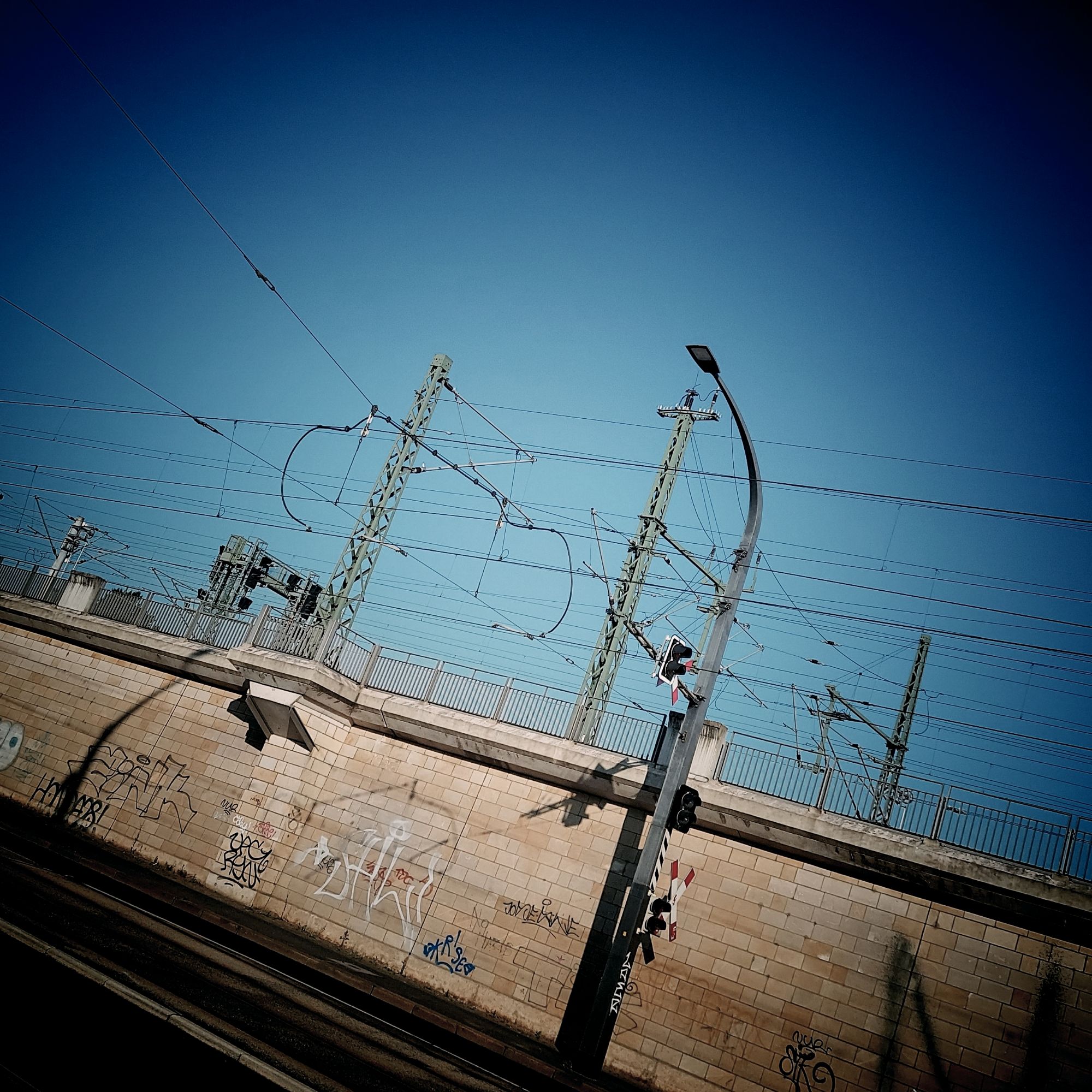 Bahntrasse, Oberleitungen und Straßenlaternen, schräg vor blassblauem Himmel.