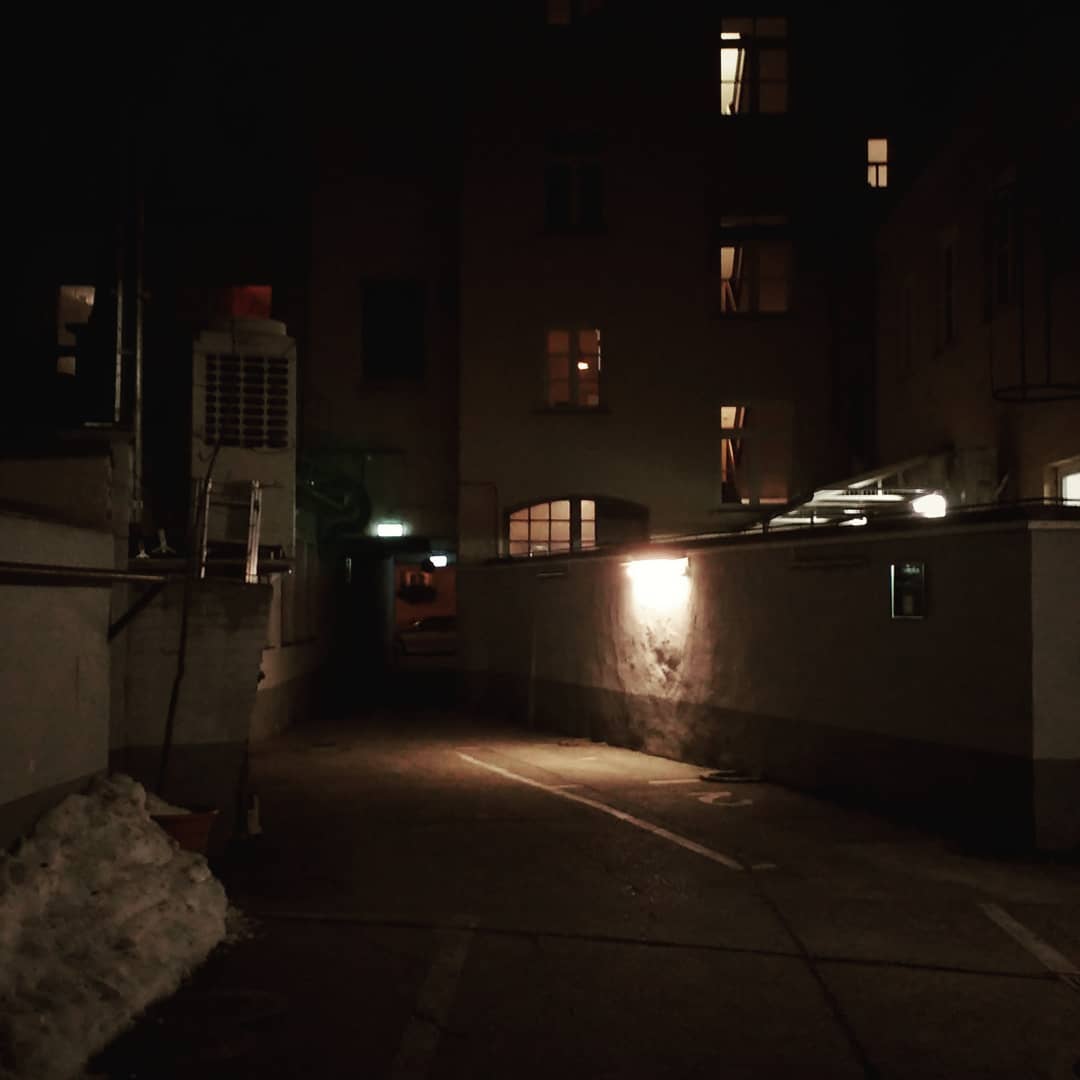 A narrow backyard with empty parking lots, walls, snow. There are a few windows alight, but most of the setting is night.